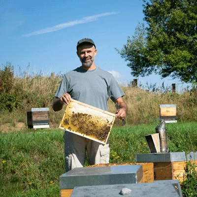 Hier siehst man der Imker Carsten Lehmann von der Imkerei Bienchenhonig mit einer Brutwabe in der Hand vor einer Bienenbeute, ohne Schleier. Hier siehst man der Imker Carsten Lehmann von der Imkerei Bienchenhonig mit einer Brutwabe in der Hand vor einer Bienenbeute, ohne Schleier.